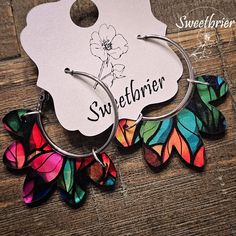 two pairs of colorful butterfly hoop earrings sitting on top of a wooden table next to a tag