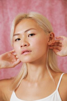 a woman with long blonde hair wearing a white tank top and holding her ear to the side