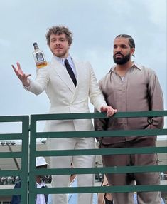 two men in white suits stand on a balcony with one holding a bottle of whiskey