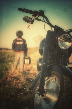 a man standing next to a motorcycle on top of a grass covered field with a person in the background