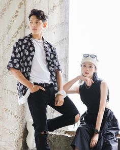 a man and woman sitting next to each other in front of a stone wall wearing sunglasses