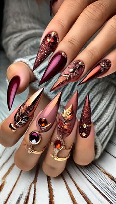 How to Create This Viral Autumn Glam Stiletto Nail Design:  Prep the Nails: Shape your nails into sharp stilettos and apply a base coat. Cure under a UV/LED lamp.  Apply Base Colors: Use deep burgundy, burnt orange, and warm taupe shades across different nails. Apply two coats for full opacity, curing each layer.  Add Glitter and Foil Accents: Apply fine glitter on select nails for a sparkling effect. Add metallic leaf foil accents on others, pressing them onto the tacky layer of polish for a smooth application.  Leaf & Gem Detailing: Use a fine nail art brush and metallic gel polish to draw leaf designs on some nails. Attach gemstones and small charms with nail glue near the cuticle area for added glamour.  Top Coat & Cure: Seal the design with a high-shine top coat Fall Nails Stilleto Shape, Burgundy Stiletto Nails, Fall Stiletto Nails Design, Burnt Orange Fall Nails, Stiletto Nail Design, Different Nails, Shape Your Nails, Draw Leaf