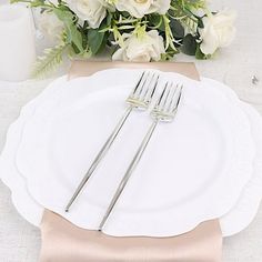 a white plate topped with two silver forks and a vase filled with flowers behind it