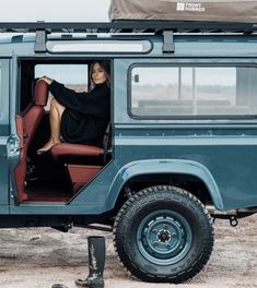 a woman sitting in the driver's seat of a blue jeep parked on top of a dirt field