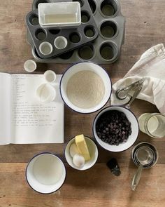 an open book sitting on top of a wooden table next to bowls and measuring cups