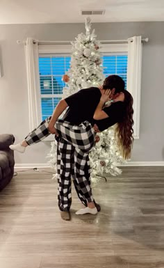 a woman in black and white checkered pants kissing a man's face next to a christmas tree