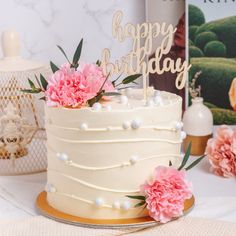 a white cake with pink carnations and a happy birthday sign on top is sitting on a table