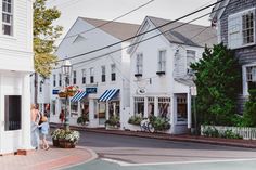 two people walking down the street in front of white buildings with shops on each side