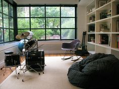 a room with a drum set, stools and bookshelves in front of a large window