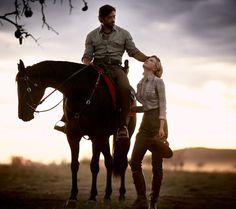 a man standing next to a woman on top of a horse near a tree at sunset