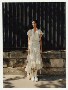 a woman standing in front of a stone wall wearing white boots and a ruffled dress