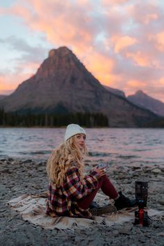 a woman sitting on top of a blanket next to a lake with mountains in the background