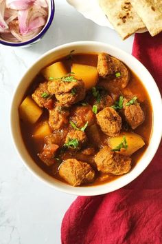 a white bowl filled with meat and potatoes next to pita bread on a red towel