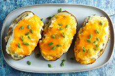 three baked potatoes with cheese and chives on a white plate, sitting on a blue surface