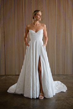 a woman standing in front of a wooden wall wearing a white dress with high slit