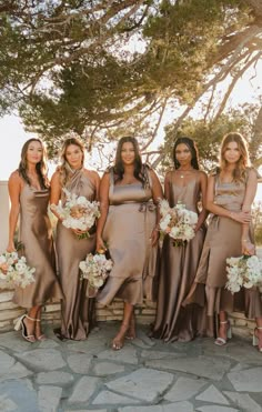 a group of women standing next to each other in front of a stone wall holding bouquets