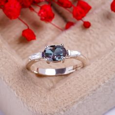 an engagement ring with a blue diamond and two white diamonds on it, sitting in front of red flowers