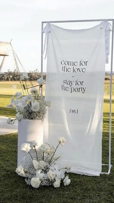 a white banner with some flowers in it on the grass next to a sign that says, come for the love of god stay for the party