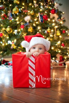 a baby wearing a santa hat sitting in front of a christmas tree holding a red bag