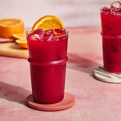 two glasses filled with red liquid and orange slices on a pink tablecloth next to sliced oranges