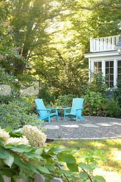 two blue chairs sitting on top of a gravel patio next to a white house and trees