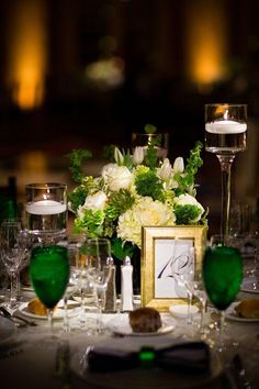 the table is set with white flowers and green wine glasses, silverware and candles