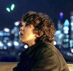 a man with curly hair sitting on top of a wooden bench in front of a cityscape