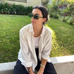 a woman sitting on the ground wearing sunglasses and a white shirt with black pants, is looking off to the side