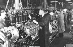 two men working on an engine in a factory with other people looking at the machines