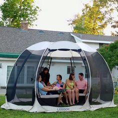 four people sitting in a large tent on the grass near a house with a white roof