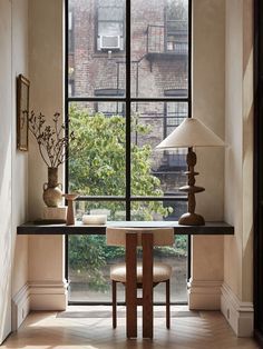 a room with a table, chair and lamp in front of a large window that looks out onto the street