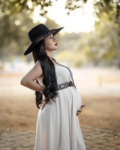 a pregnant woman wearing a black hat and white dress standing in front of a tree