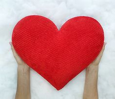 two hands holding a red heart shaped pillow in the middle of white furnishing