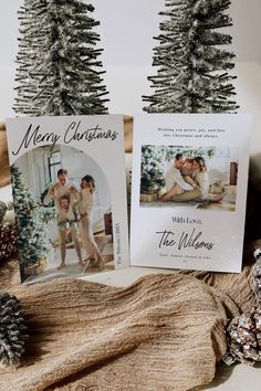 two christmas cards sitting on top of a table next to pine cones and fir trees