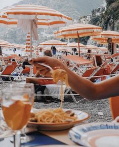 an outdoor dining area with orange umbrellas and people eating spaghetti on the beach in front of them