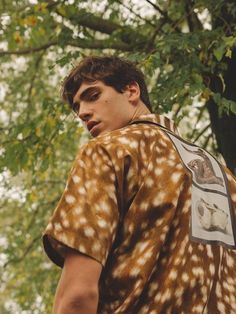 a young man wearing a brown and white deer print shirt standing next to a tree