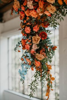 an arrangement of flowers and greenery hanging from the ceiling in front of a window