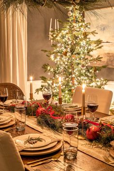 a dining room table set for christmas dinner with candles and plates on it, next to a decorated christmas tree