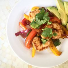 a white plate topped with shrimp and veggies next to sliced avocado