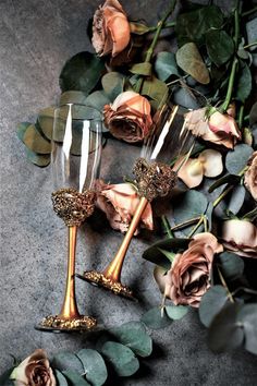 two champagne flutes with gold glittered handles and rose petals on a gray surface surrounded by greenery