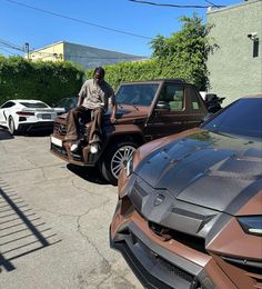 a man sitting on the hood of a brown sports utility truck parked next to other cars