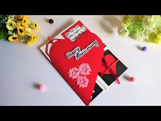 a red heart shaped card sitting on top of a table next to flowers and confetti