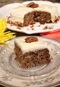 a piece of carrot cake on a glass plate next to a yellow flower with pecans in the background