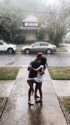 two people hugging each other in the rain on a sidewalk with cars parked behind them