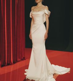 a woman standing in front of a red curtain wearing a white dress with one shoulder