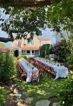 a long table set up in the yard for an outdoor dinner party with white linens and wooden chairs