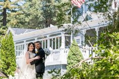 a bride and groom standing in front of a white house with an american flag on the porch