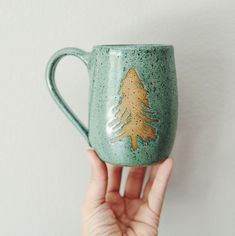 a hand holding a green ceramic mug with a gold pine tree painted on the side