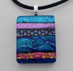 a square pendant with blue, purple and pink designs hanging from a black leather cord