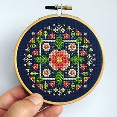 a hand is holding a cross stitched floral design on a blue background with red, yellow and green flowers